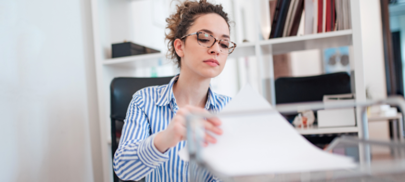 Mitarbeiterin mit Brille sortiert Papierunterlagen an einem Schreibtisch im Büro.