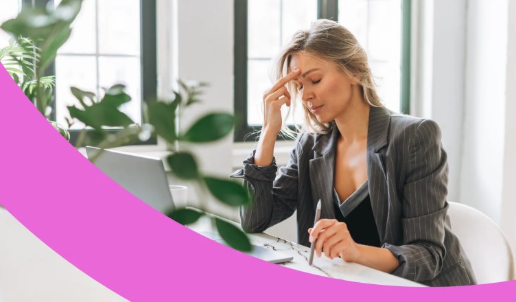 Erschöpfte junge Frau mit Boreout sitzt am Schreibtisch und hält sich mit geschlossenen Augen zwei Finger an die Stirn.