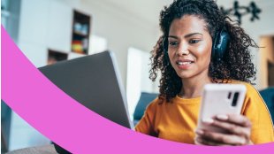 Mitarbeiterin im Homeoffice mit Kopfhörern auf den Ohren hält ein Handy und arbeitet am Laptop.