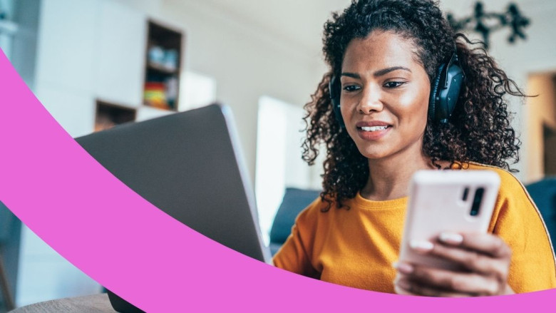 Mitarbeiterin im Homeoffice mit Kopfhörern auf den Ohren hält ein Handy und arbeitet am Laptop.