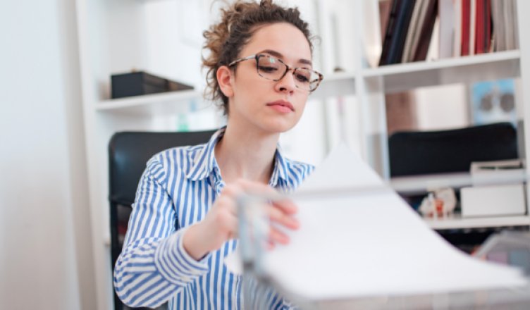 Mitarbeiterin mit Brille sortiert Papierunterlagen an einem Schreibtisch im Büro.