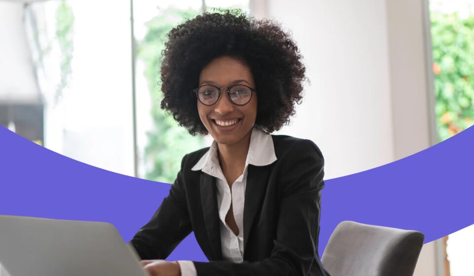 Lächelnde Frau sitzt an einem Tisch und arbeitet am Laptop.