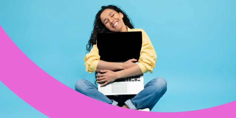 Lachende Frau sitzt im Schneidersatz auf dem Boden und umklammert ihren Laptop mit beiden Armen.