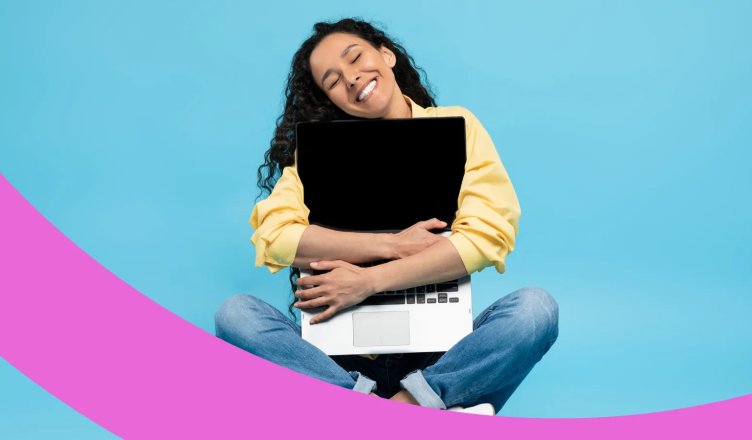 Lachende Frau sitzt im Schneidersatz auf dem Boden und umklammert ihren Laptop mit beiden Armen.
