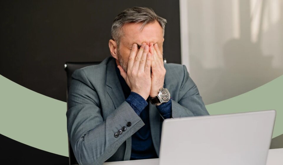 Mann sitzt am Schreibtisch und hält sich beide Hände vor das Gesicht.