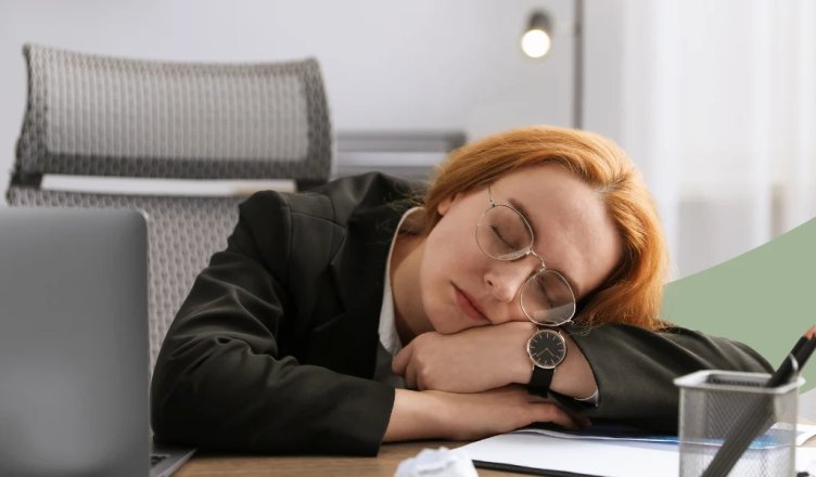 Frau schläft mit dem Kopf auf den Armen gestützt am Schreibtisch neben ihrem Laptop.