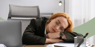 Frau schläft mit dem Kopf auf den Armen gestützt am Schreibtisch neben ihrem Laptop.