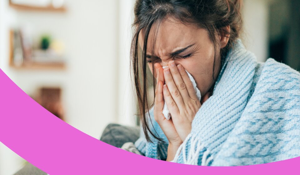 Frau sitzt mit einer Decke über den Schultern auf dem Sofa und putzt sich mit beiden Händen die Nase.