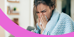 Frau sitzt mit einer Decke über den Schultern auf dem Sofa und putzt sich mit beiden Händen die Nase.